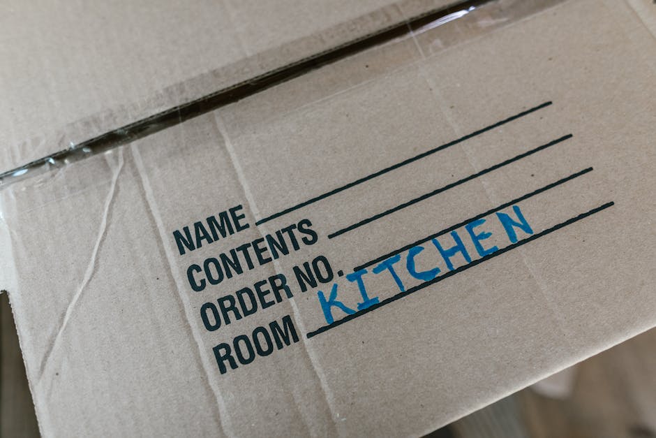 Close-up of a cardboard moving box with a label indicating contents and destination. The label shows the handwritten word 'KITCHEN' in blue ink, along with printed sections for 'name,' 'contents,' 'or
